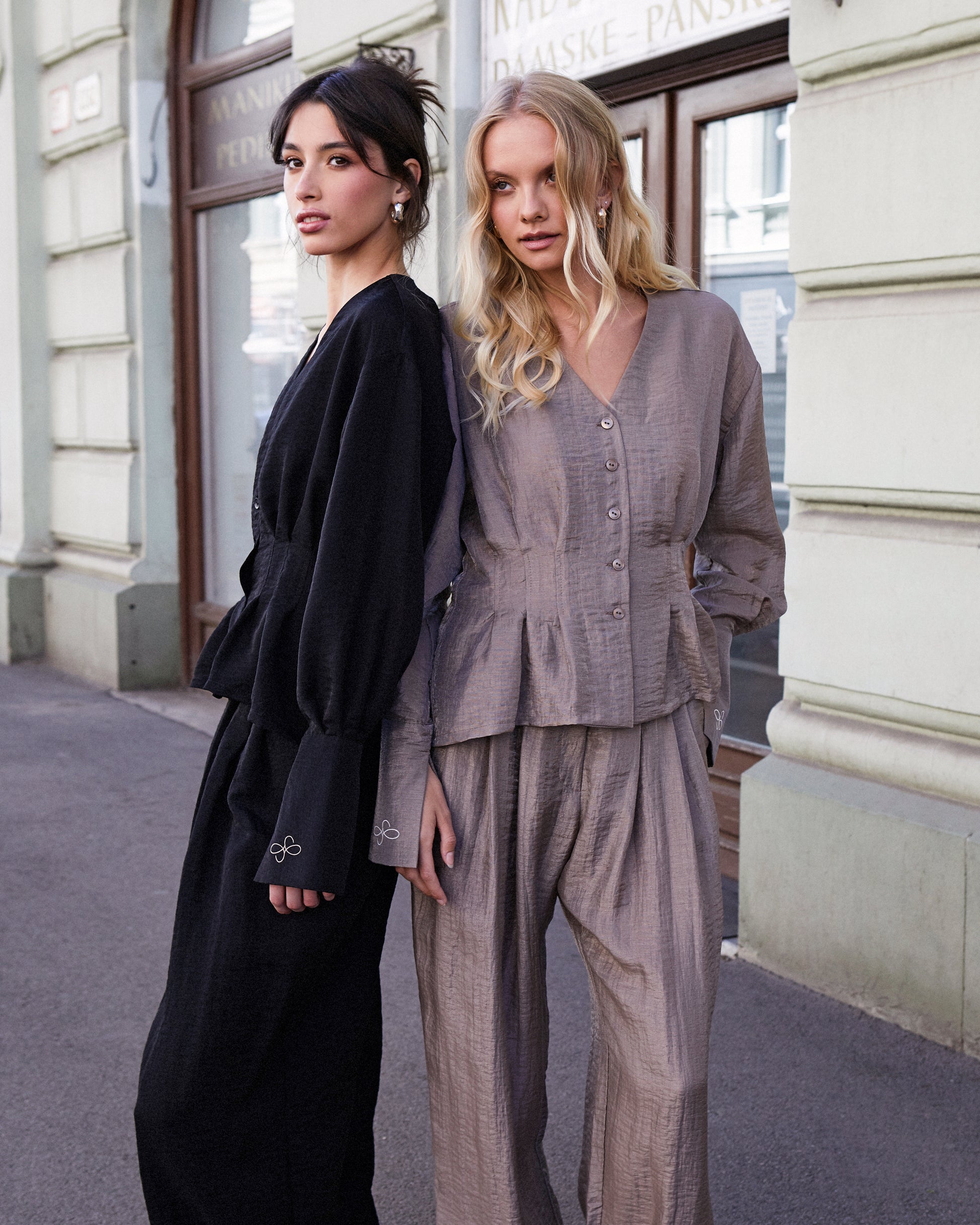 Two women standing side by side on a city street, one in a black outfit and the other in a gray outfit.